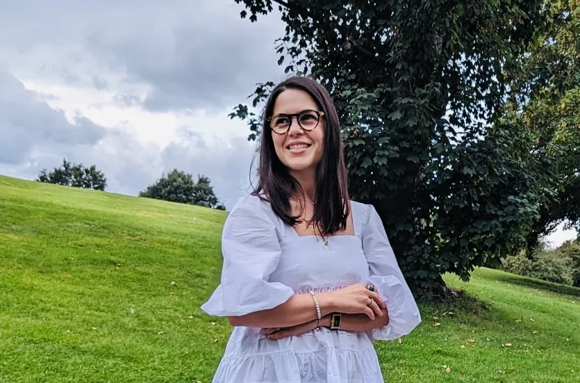 Rocío León smiling with her arms crossed, standing on a grassy hill in front of a large tree under a cloudy sky.